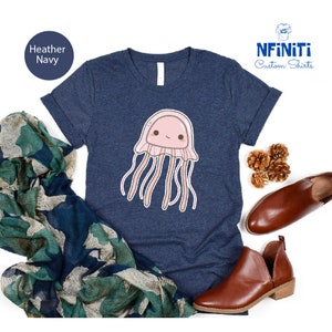 May include: Heather navy blue t-shirt with a cute cartoon jellyfish graphic. The jellyfish is pink with a white face and has long pink tentacles. The shirt has a crew neck and short sleeves. The text "NFINITI Custom Shirts" is in the top right corner of the image.