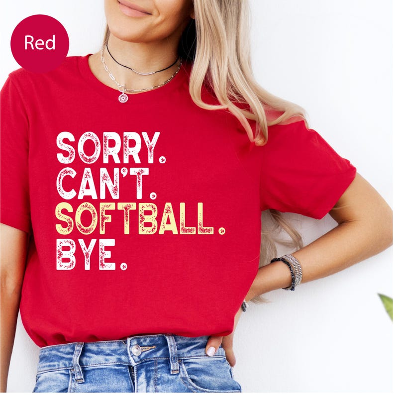 May include: A red short-sleeved t-shirt featuring white and yellow distressed-style text that reads: SORRY. CAN'T. SOFTBALL. BYE. The shirt has a crew neckline and is paired with blue denim jeans, creating a casual and sporty appearance.