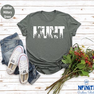 May include: A heather military green t-shirt with the word "HUNT" in white text. The letters are outlined with silhouettes of a hunter and a deer in the woods.