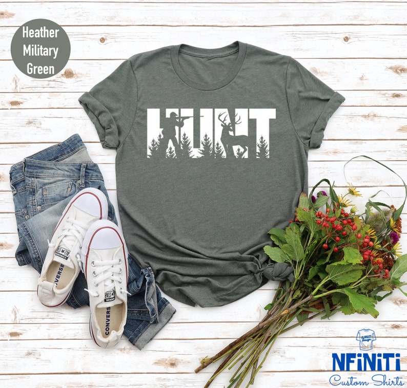 May include: A heather military green t-shirt with the word "HUNT" in white text. The letters are outlined with silhouettes of a hunter and a deer in the woods.