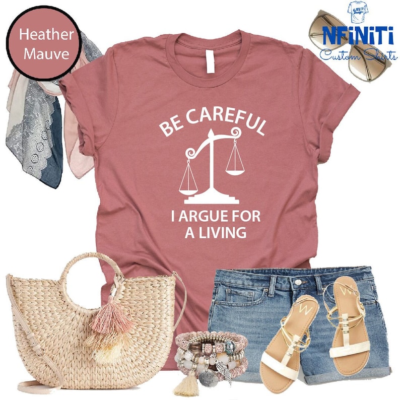 May include: Heather mauve t-shirt with the text "Be Careful I Argue For A Living" and a white scale graphic. The shirt is paired with denim shorts, a straw bag with a pink tassel, and white sandals.