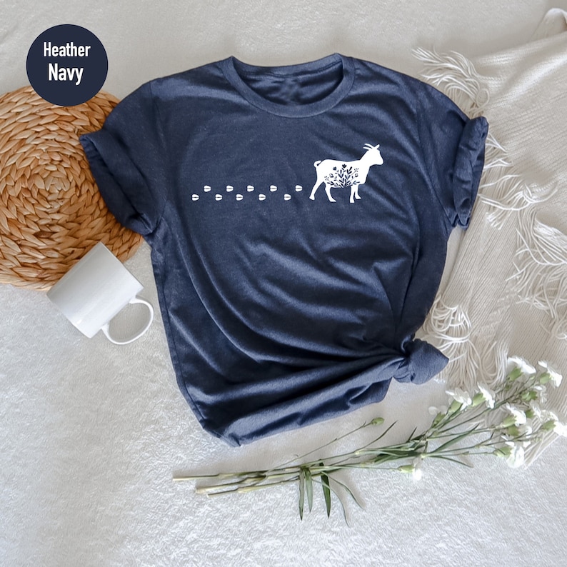 Pu&ograve; includere: Una t-shirt blu navy heather con una silhouette bianca di una capra con dettagli floreali e una scia di impronte.