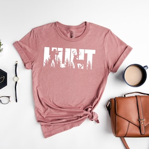 May include: A heather mauve t-shirt with the word "HUNT" in white text. The letters are made up of silhouettes of a hunter, trees, and a deer.