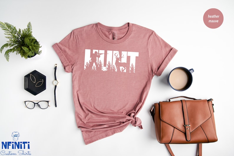 May include: A heather mauve t-shirt with the word "HUNT" in white text. The letters are made up of silhouettes of a hunter, trees, and a deer.