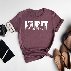 May include: A heather maroon t-shirt with the word "HUNT" in white text. The letters are made up of silhouettes of a hunter, a deer, and trees.