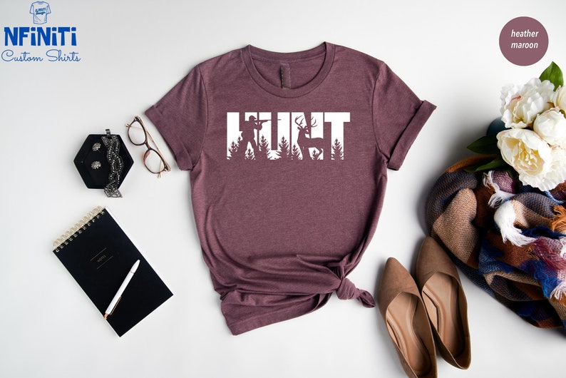May include: A heather maroon t-shirt with the word "HUNT" in white text. The letters are made up of silhouettes of a hunter, a deer, and trees.