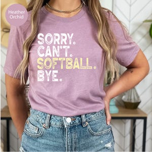 May include: A heather orchid short-sleeve t-shirt featuring the white and yellow text SORRY. CAN'T. SOFTBALL. BYE. printed on the front. The shirt has a crew neckline and a standard fit, paired with blue denim jeans.