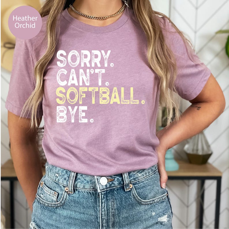 May include: A heather orchid short-sleeve t-shirt featuring the white and yellow text SORRY. CAN'T. SOFTBALL. BYE. printed on the front. The shirt has a crew neckline and a standard fit, paired with blue denim jeans.
