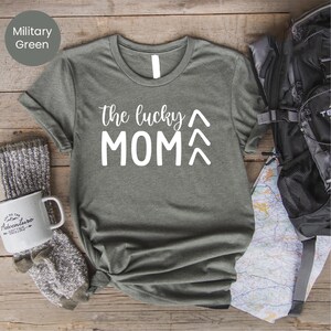 May include: Military green t-shirt with the text "the lucky MOM" in white. The shirt is styled with a backpack, a mug that says "Adventure Begins", and a pair of gray socks. The shirt is made of a soft material.