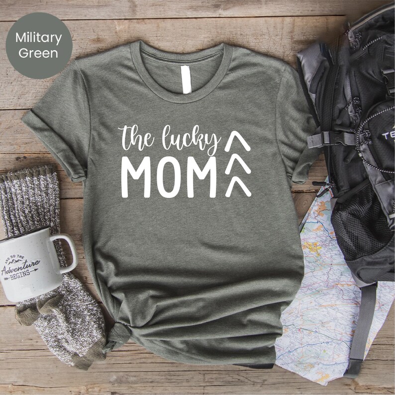 May include: Military green t-shirt with the text "the lucky MOM" in white. The shirt is styled with a backpack, a mug that says "Adventure Begins", and a pair of gray socks. The shirt is made of a soft material.