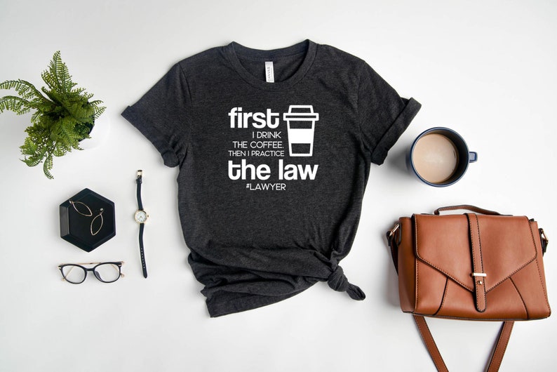 以下が含まれることがあります： 白い文字で「first I drink the coffee then I practice the law #LAWYER」と書かれた濃いグレーのTシャツ。Tシャツは裾が結ばれており、テキストの横にコーヒーカップのアイコンがあります。