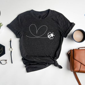 Peut inclure: T-shirt en gris fonc&eacute; chin&eacute; avec un motif d'abeille blanche volant en forme de c&oelig;ur. L'abeille est dessin&eacute;e en blanc et a des rayures noires. Le t-shirt est pos&eacute; &agrave; plat sur une surface blanche avec une montre, des lunettes, des boucles d'oreilles, une tasse &agrave; caf&eacute; et un sac &agrave; main en cuir marron.