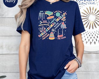May include: Navy blue t-shirt with a colourful graphic design featuring the words "Occupational Therapy" and related terms such as "Play" and "Fine Motor." The design includes illustrations of therapy tools. The shirt has a crew neck.