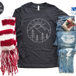 K&ouml;nnte beinhalten: Langarm-T-Shirt in Dunkelgrau meliert mit einem wei&szlig;en Umriss einer Bergkette, Sonne und B&auml;umen in einem Kreis. Das Shirt liegt flach auf einem wei&szlig;en Hintergrund mit einem rot-wei&szlig; gestreiften Schal und einer blauen Jeans.