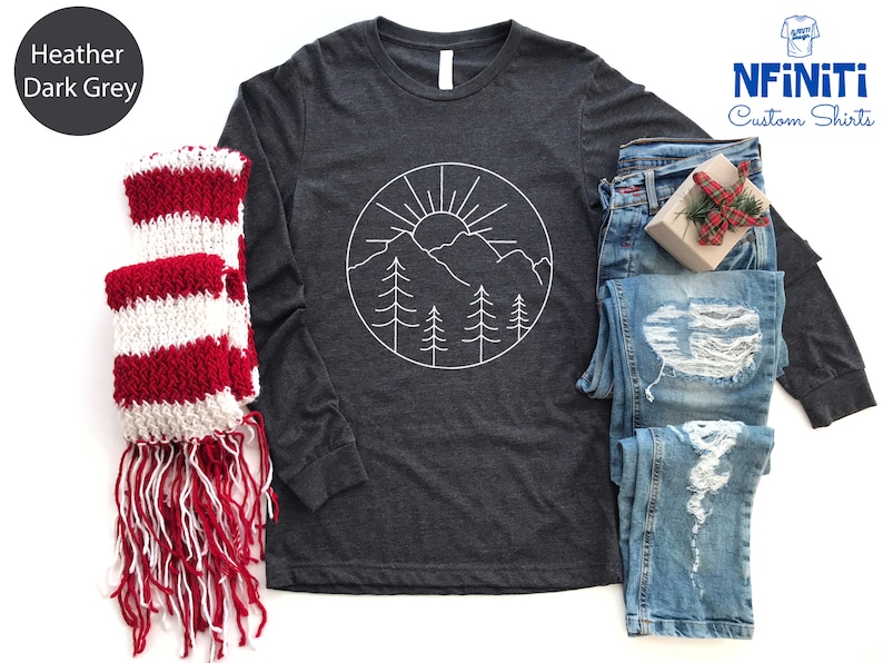 K&ouml;nnte beinhalten: Langarm-T-Shirt in Dunkelgrau meliert mit einem wei&szlig;en Umriss einer Bergkette, Sonne und B&auml;umen in einem Kreis. Das Shirt liegt flach auf einem wei&szlig;en Hintergrund mit einem rot-wei&szlig; gestreiften Schal und einer blauen Jeans.