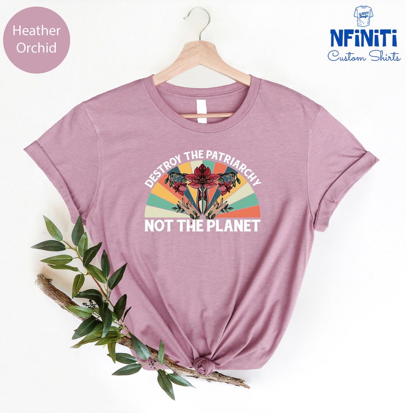 May include: A heather orchid t-shirt with a vintage-style graphic that says "Destroy the Patriarchy Not the Planet". The graphic features a stylized uterus with flowers and a rainbow.