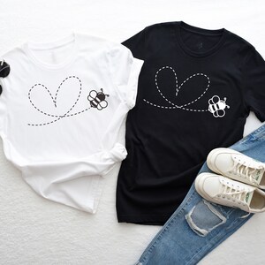 Peut inclure: Deux t-shirts avec une abeille volant en ligne pointill&eacute;e qui forme un c&oelig;ur. Les t-shirts sont blanc et noir. Le t-shirt blanc est &agrave; gauche et le t-shirt noir est &agrave; droite. Les t-shirts sont dispos&eacute;s sur une surface blanche avec un jean et des baskets blanches.