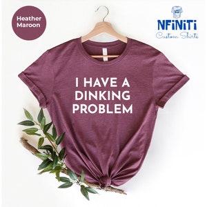 Peut inclure: Un t-shirt en marron chiné avec le texte "I HAVE A DINKING PROBLEM" imprimé en blanc.