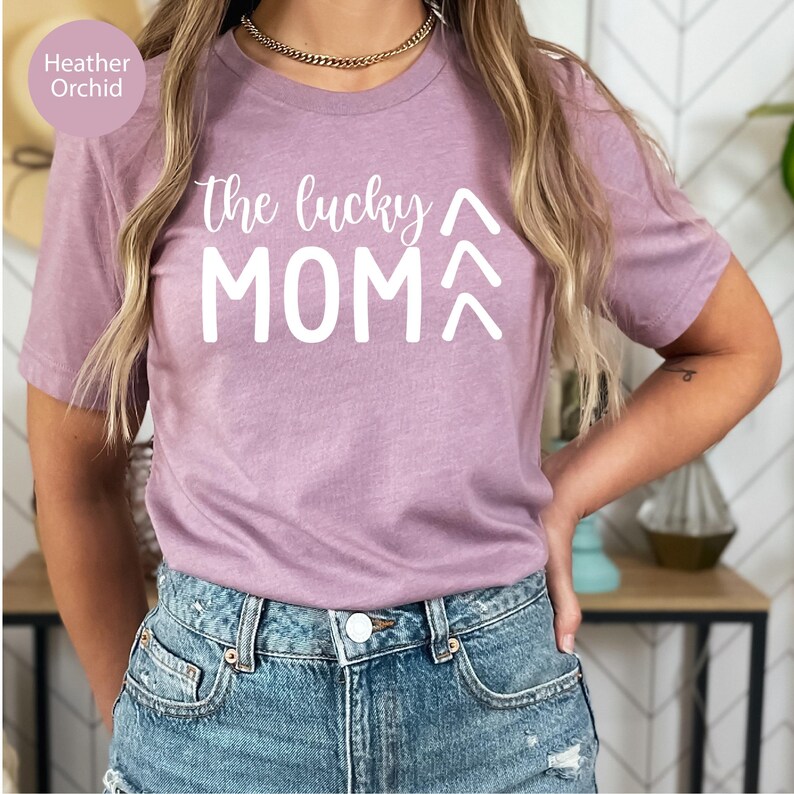 May include: Heather orchid-colored t-shirt with the text "the lucky MOM" in white. The shirt has a crew neck and short sleeves. The model is wearing blue denim shorts. The shirt is made of a soft, comfortable material.