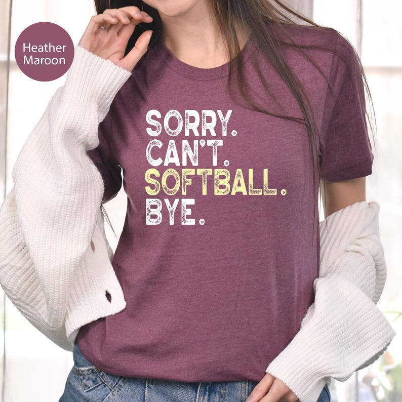 May include: A heather maroon short-sleeve t-shirt featuring white and yellow distressed text that reads SORRY. CAN'T. SOFTBALL. BYE. The shirt is worn with a white long-sleeve cardigan and blue denim jeans, showcasing a casual layered look.