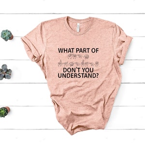 May include: A peach-colored t-shirt with the text "WHAT PART OF DON'T YOU UNDERSTAND?" in black. The text is accompanied by a row of hand signs in black, representing the word "deaf" in American Sign Language.