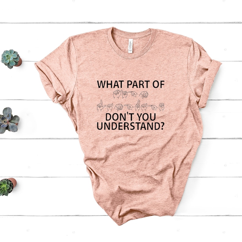 May include: A peach-colored t-shirt with the text "WHAT PART OF DON'T YOU UNDERSTAND?" in black. The text is accompanied by a row of hand signs in black, representing the word "deaf" in American Sign Language.