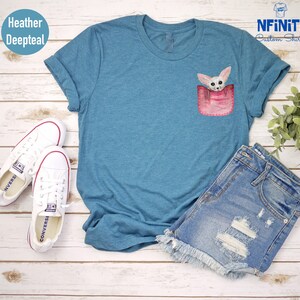 May include: Deep teal t-shirt with a pocket design featuring a fennec fox illustration. The shirt is styled with denim shorts and white Converse trainers. The sleeves are rolled up. The shirt is made of a soft material.