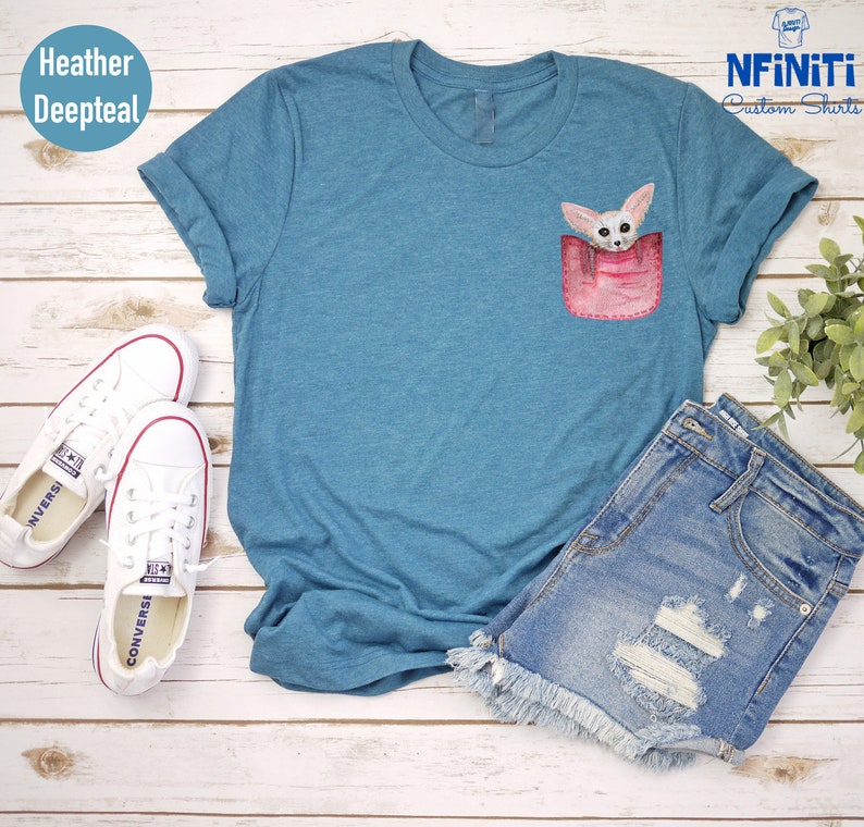 May include: Deep teal t-shirt with a pocket design featuring a fennec fox illustration. The shirt is styled with denim shorts and white Converse trainers. The sleeves are rolled up. The shirt is made of a soft material.