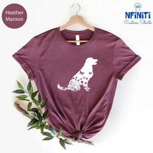 May include: Heather maroon t-shirt with a white silhouette of a dog made up of flowers and butterflies. The shirt is on a hanger and has a white background.