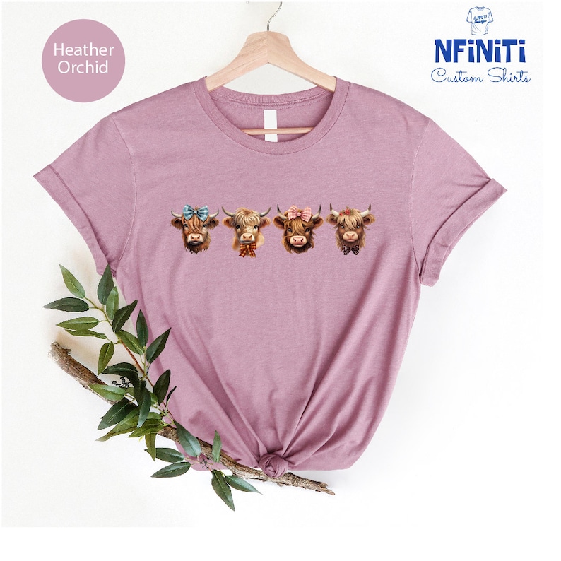 Puede incluir: Una camiseta de color orqu&iacute;dea en heather con una fila de cuatro caras de vacas Highland que llevan lazos y cintas. Las vacas son marrones y blancas con narices negras. La camiseta est&aacute; en una percha y tiene un fondo blanco.