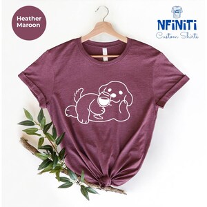 May include: Heather maroon t-shirt with a white graphic of a dog holding a glass of wine. The shirt is on a hanger and has a white label that says "NFINITI Custom Shirts".
