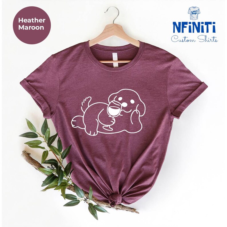 May include: Heather maroon t-shirt with a white graphic of a dog holding a glass of wine. The shirt is on a hanger and has a white label that says "NFINITI Custom Shirts".
