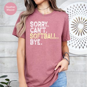 May include: Mauve heather t-shirt featuring the text "SORRY. CAN'T. SOFTBALL. BYE." in white and yellow. The top is a short-sleeved crew neck style.