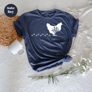 May include: A heather navy blue t-shirt with a white silhouette of a chicken with floral details and a trail of chicken footprints.