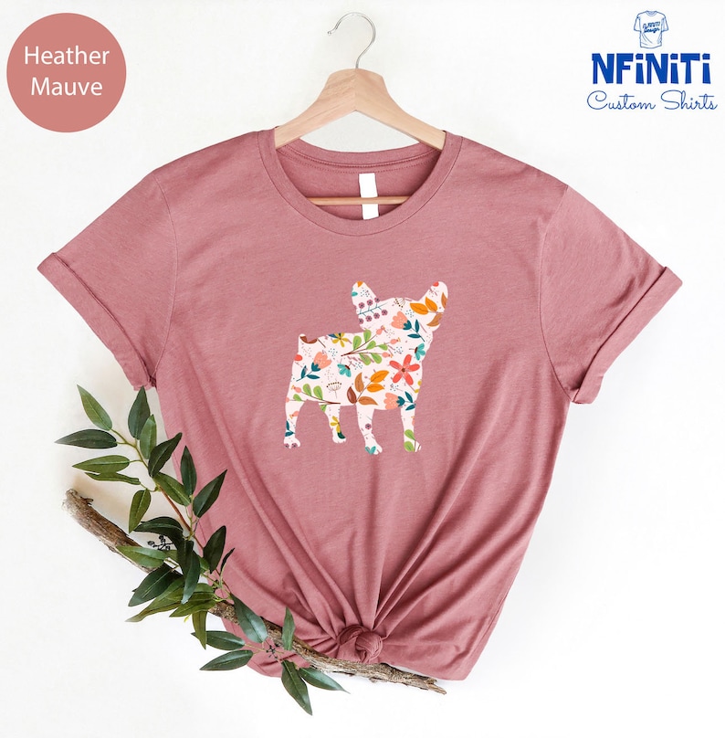 Peut inclure: Un t-shirt en mauve chin&eacute; avec un imprim&eacute; floral en forme de silhouette de bouledogue fran&ccedil;ais. Le t-shirt est sur un cintre et a un fond blanc.