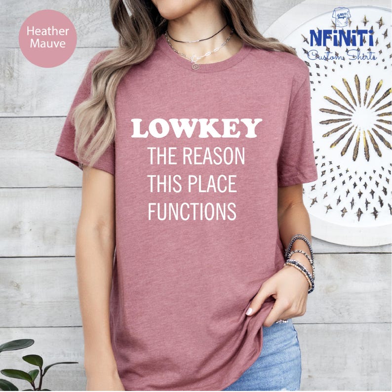 A woman wearing a pink t-shirt with the text "LOWKEY THE REASON THIS PLACE FUNCTIONS" printed on it.