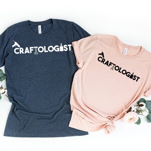 May include: Two t-shirts with the text "CRAFTOLOGIST" printed on them. The shirts are in different colors, one is navy blue and the other is pink. The text is in black on the pink shirt and white on the navy blue shirt. The text is accompanied by a graphic of a glue bottle, scissors, and a paintbrush.