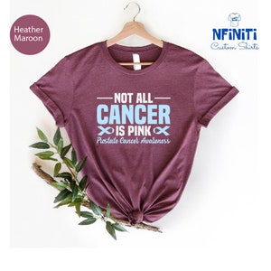 May include: Heather maroon t-shirt with the text "Not all cancer is pink" and a blue ribbon symbol. The text "Prostate Cancer Awareness" is below the ribbon.