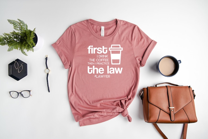 以下が含まれることがあります： 「first I drink the coffee then I practice the law #lawyer」というテキストと白いコーヒーカップのグラフィックが描かれたピンクのTシャツです。