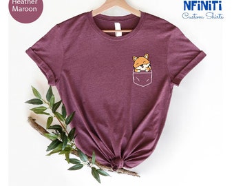Puede incluir: Camiseta de color marrón heather con un bolsillo que presenta un zorro de dibujos animados asomándose. La camiseta está sobre un fondo blanco con una rama de hojas verdes.