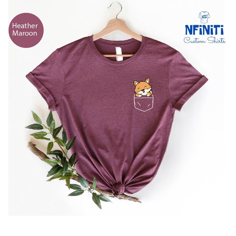 May include: Heather maroon t-shirt with a pocket featuring a cartoon fox peeking out. The shirt is on a white background with a branch of green leaves.