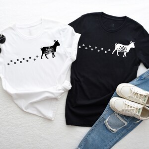 Pu&ograve; includere: Due t-shirt con una silhouette nera di una capra con dettagli floreali e una scia di impronte. Le t-shirt sono bianche e nere. La t-shirt bianca &egrave; a sinistra e la t-shirt nera &egrave; a destra. Un paio di scarpe da ginnastica bianche e un paio di jeans blu sono nell'angolo in basso a destra dell'immagine.