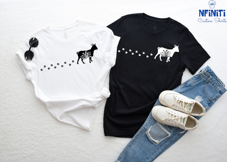 Pu&ograve; includere: Due t-shirt con una silhouette nera di una capra con dettagli floreali e una scia di impronte. Le t-shirt sono bianche e nere. La t-shirt bianca &egrave; a sinistra e la t-shirt nera &egrave; a destra. Un paio di scarpe da ginnastica bianche e un paio di jeans blu sono nell'angolo in basso a destra dell'immagine.