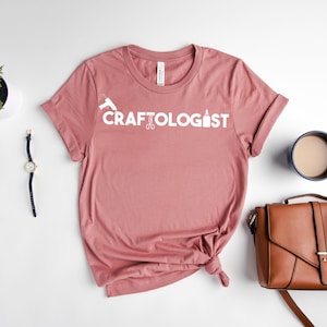 May include: A dusty rose colored t-shirt with the text "CRAFTOLOGIST" in white lettering. The text is accompanied by a graphic of a glue bottle, scissors, and a hot glue gun.