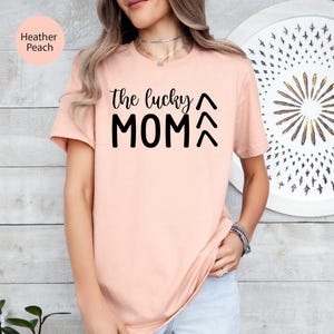 May include: Heather peach-colored t-shirt with the text "the lucky MOM" in black. The shirt has short sleeves and a crew neck. The text is in a bold, sans-serif font.