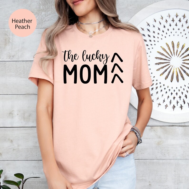 May include: Heather peach-colored t-shirt with the text "the lucky MOM" in black. The shirt has short sleeves and a crew neck. The text is in a bold, sans-serif font.