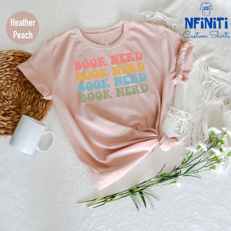 May include: A heather peach t-shirt with the text "BOOK NERD" repeated four times in a rainbow color scheme. The text is in a retro font style.