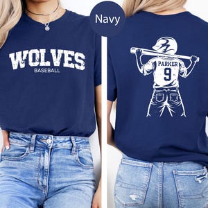 May include: Navy blue baseball t-shirt. The front features the word "WOLVES" above "BASEBALL" in white distressed lettering. The back displays a white illustration of a baseball player holding a bat, with the name "PARKER" and the number "9".