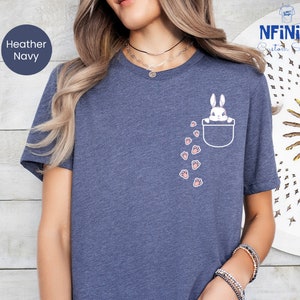 May include: A heather navy blue t-shirt with a white pocket featuring a white bunny peeking out and pink paw prints trailing behind it.