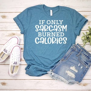 May include: A teal blue t-shirt with the text "If only sarcasm burned calories" in white lettering. The shirt is laid out on a wooden surface with a pair of white trainers and denim shorts.